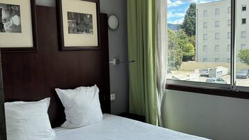 Une chambre d’hôtel avec un lit, des tableaux encadrés accrochés au mur, une fenêtre avec des rideaux et une vue sur les bâtiments à l’extérieur.