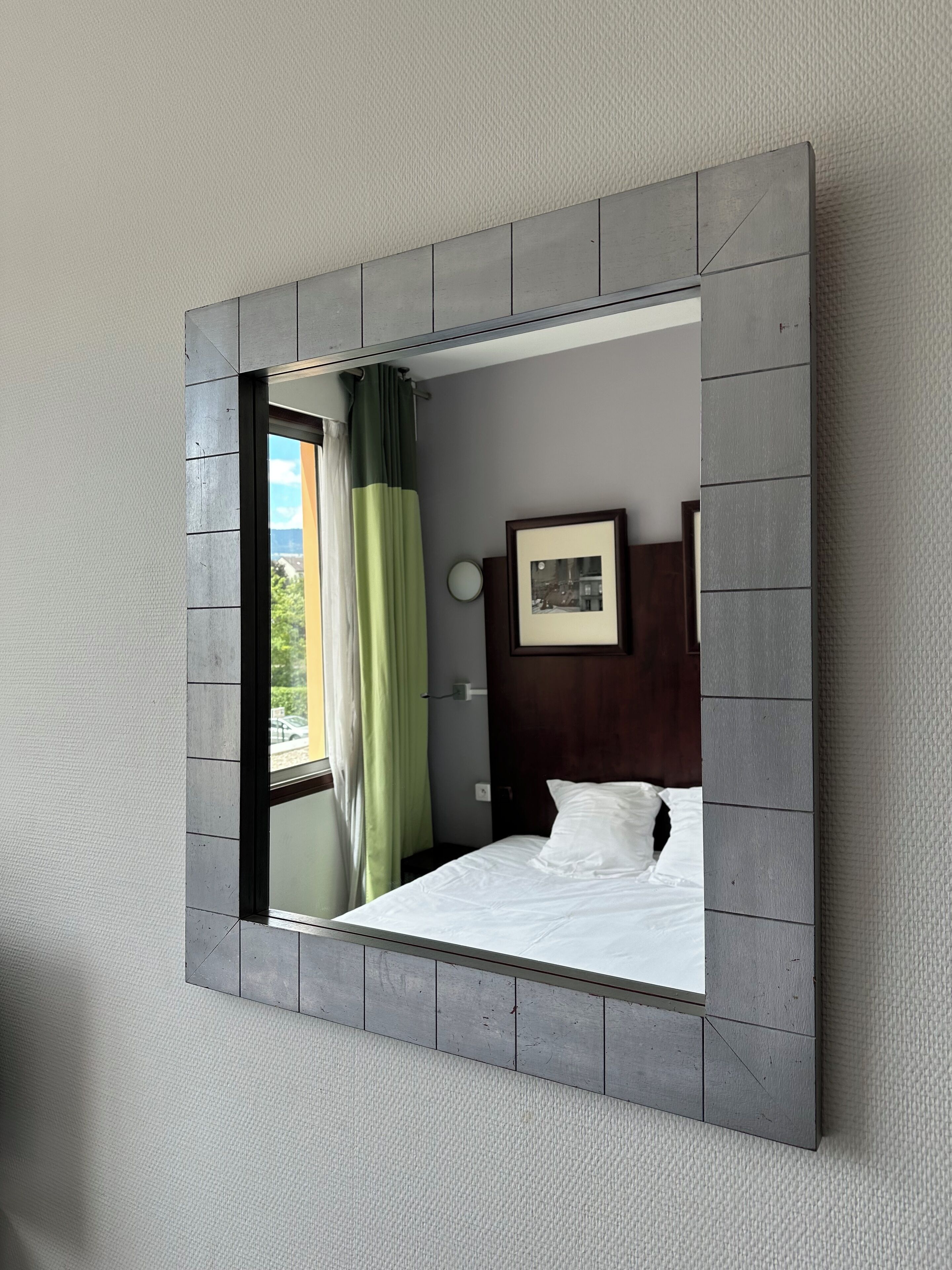 Habitación de hotel con un espejo grande, una cama con ropa blanca, un cabecero de madera y un cuadro en la pared.