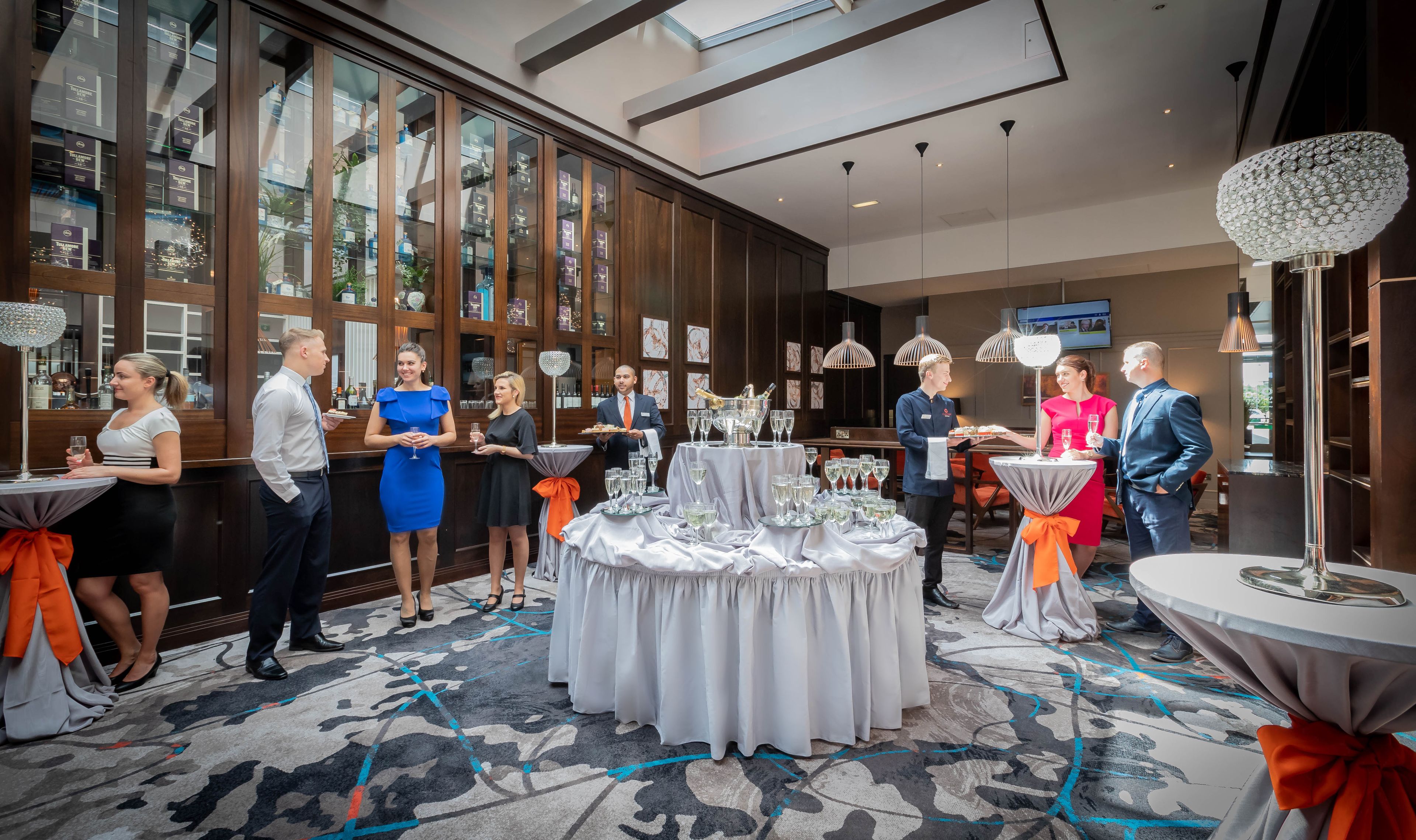 A formal event in a spacious room with large windows, a central table with a white tablecloth, and a chandelier.