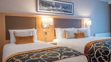 A hotel room with two beds, each with a headboard, white bedding, and decorative pillows. There are wall-mounted lamps on either side of the beds.