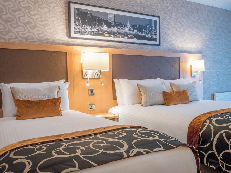 A hotel room with two beds, each with a headboard, white bedding, and decorative pillows. There are wall-mounted lamps on either side of the beds.