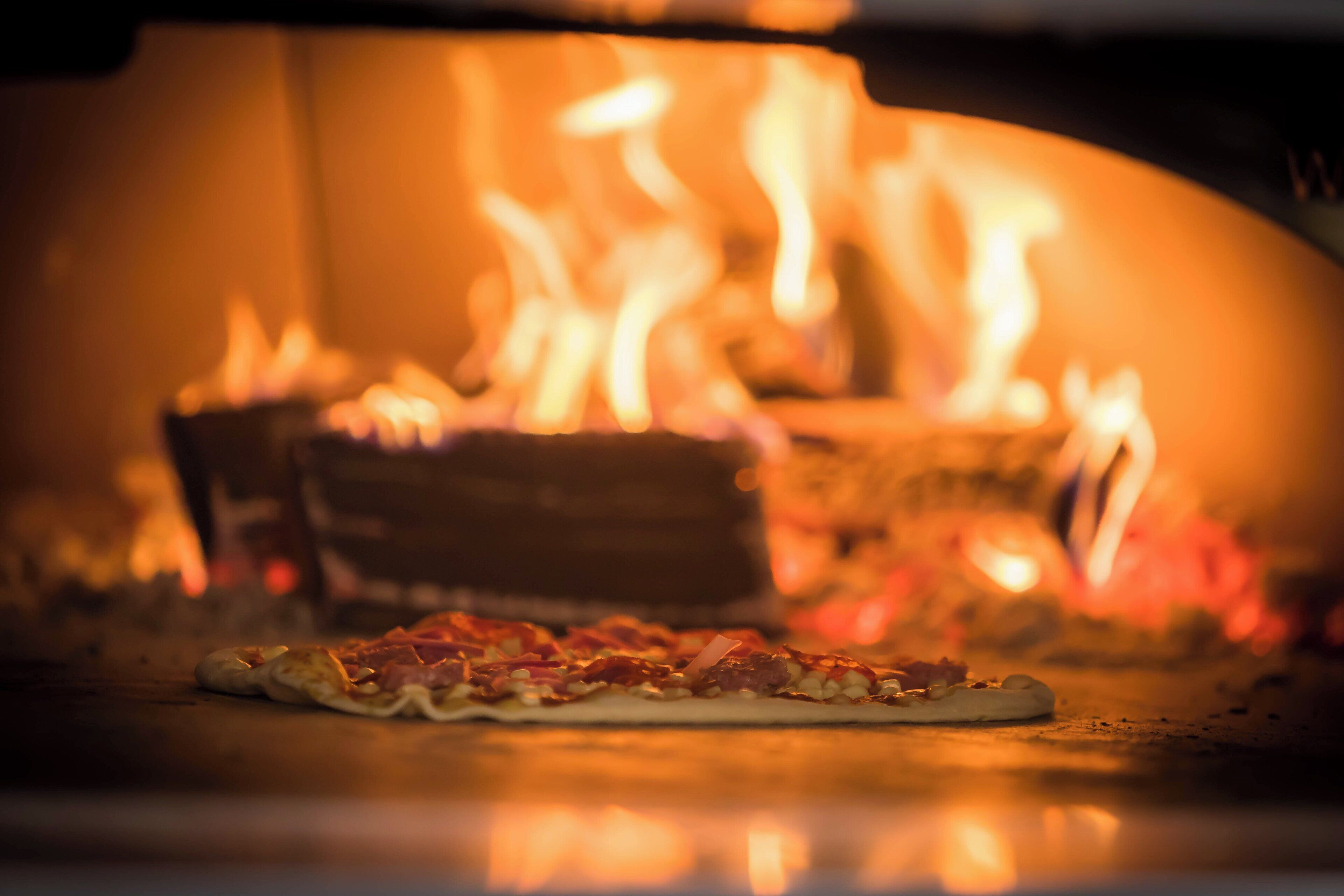 A pizza in a wood-fired oven with flames visible in the background.