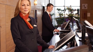 A reception area with a receptionist at a desk, a man in the background, and a view of the outdoors through large windows.