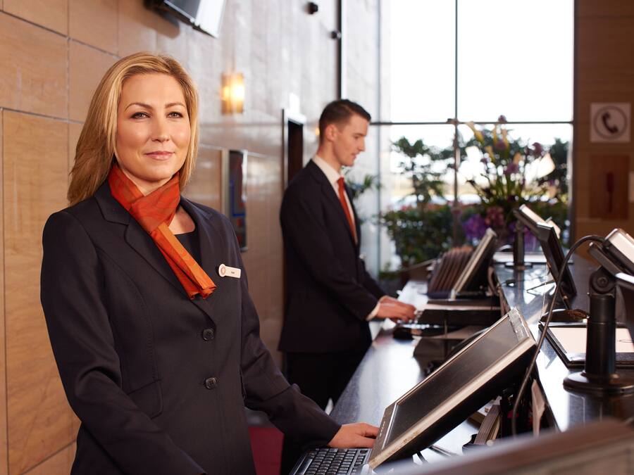 A reception area with a receptionist at a desk, a man in the background, and a view of the outdoors through large windows.