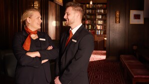 Two people in a formal setting with a bookshelf and a patterned carpet.