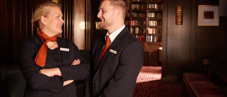 Two people in a formal setting with a bookshelf and a patterned carpet.