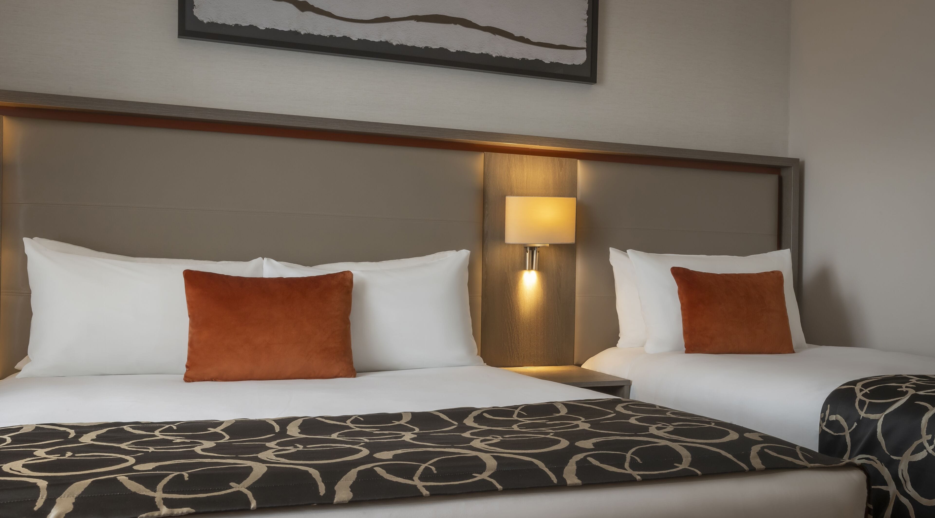 A hotel room with two beds, each with white bedding and brown pillows. There is a nightstand with a lamp between the beds. The room has a modern design with a wooden headboard and a framed artwork above the beds.