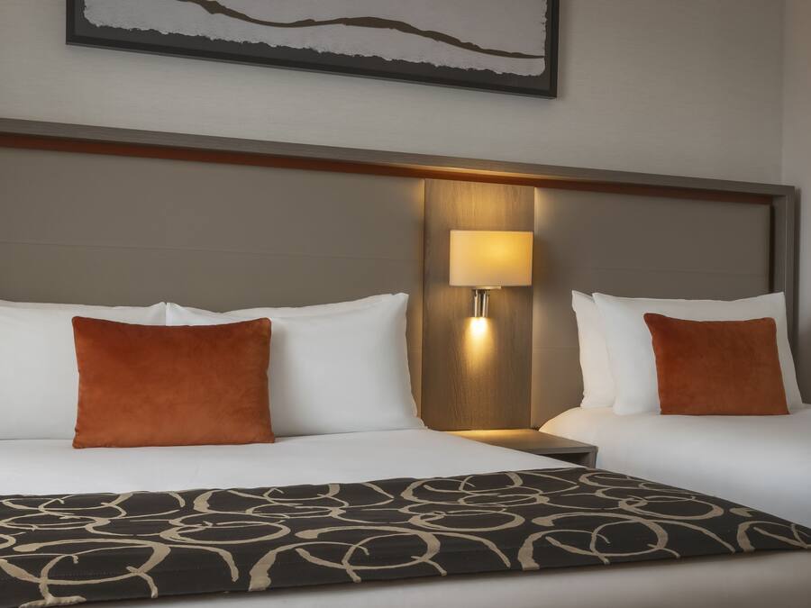 A hotel room with two beds, each with white bedding and brown pillows. There is a nightstand with a lamp between the beds. The room has a modern design with a wooden headboard and a framed artwork above the beds.