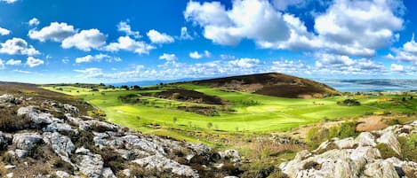 A panoramic view of a golf course with rolling hills, a large hill in the center, and a body of water in the distance.