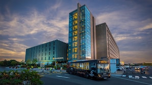 A modern hotel with a glass facade, a shuttle bus in front, and a parking lot.