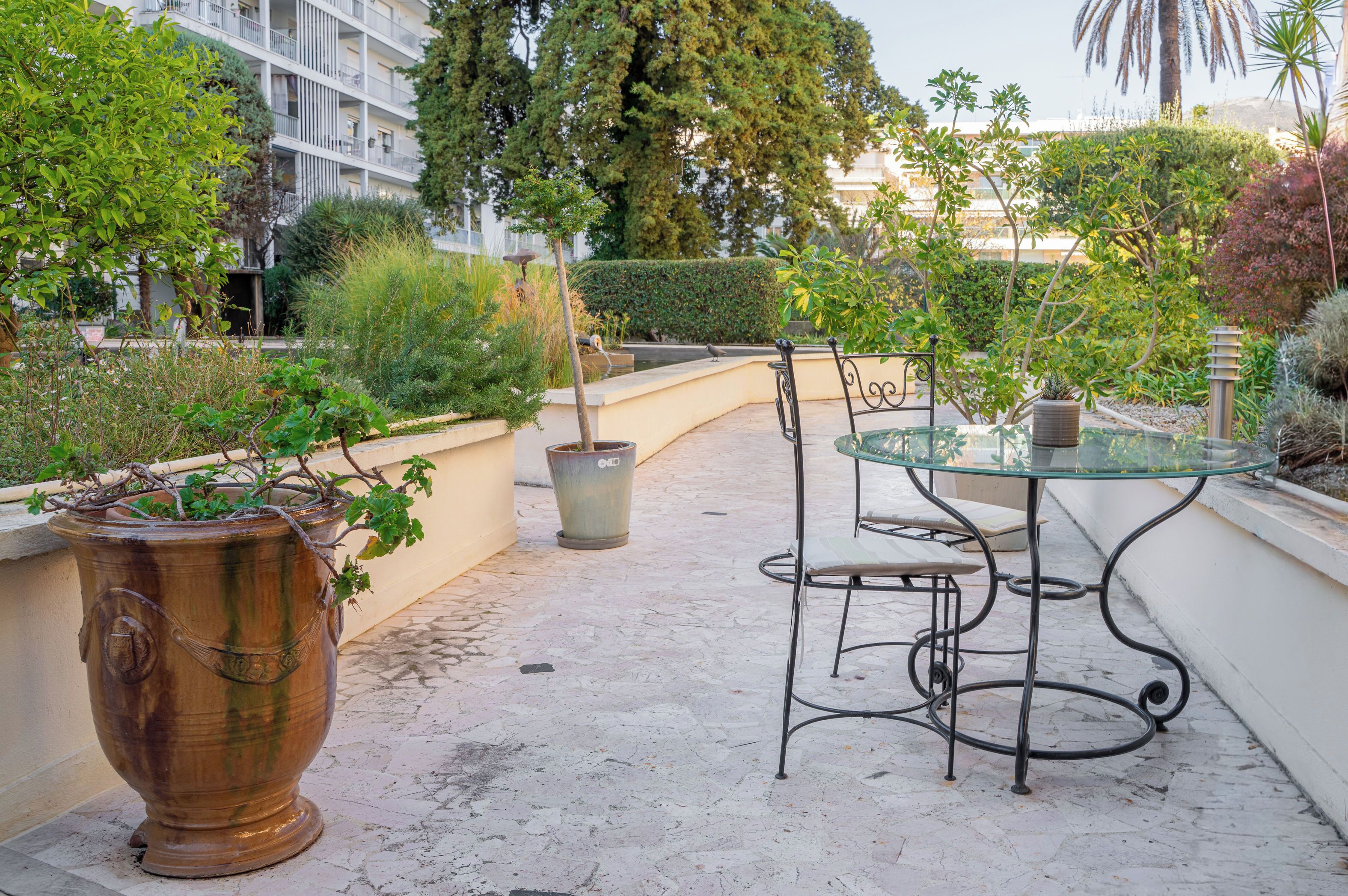 A patio with a glass table and metal chairs, surrounded by potted plants and a large urn.