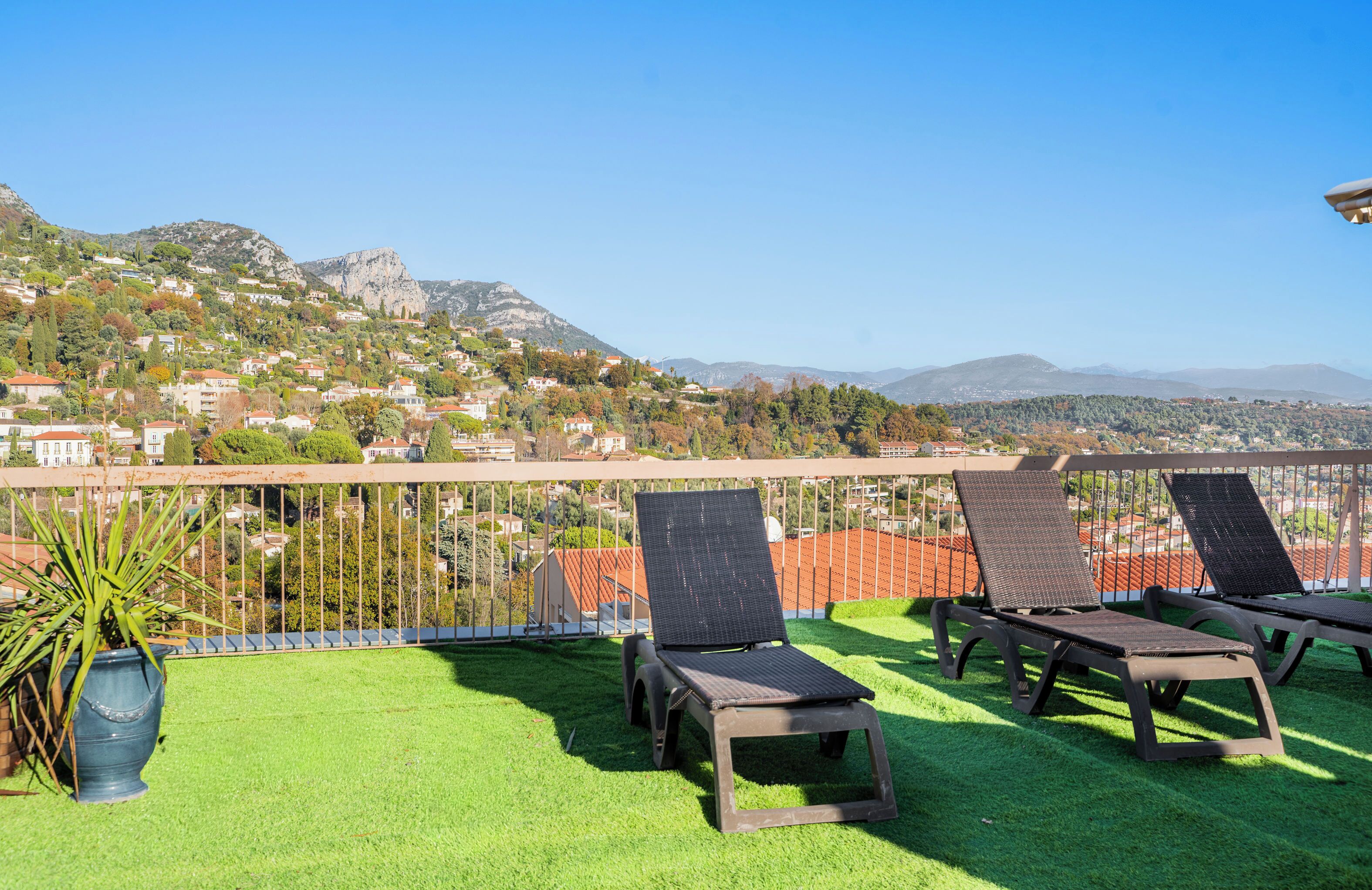 A rooftop terrace with three lounge chairs, a potted plant, and a view of a hillside town.
