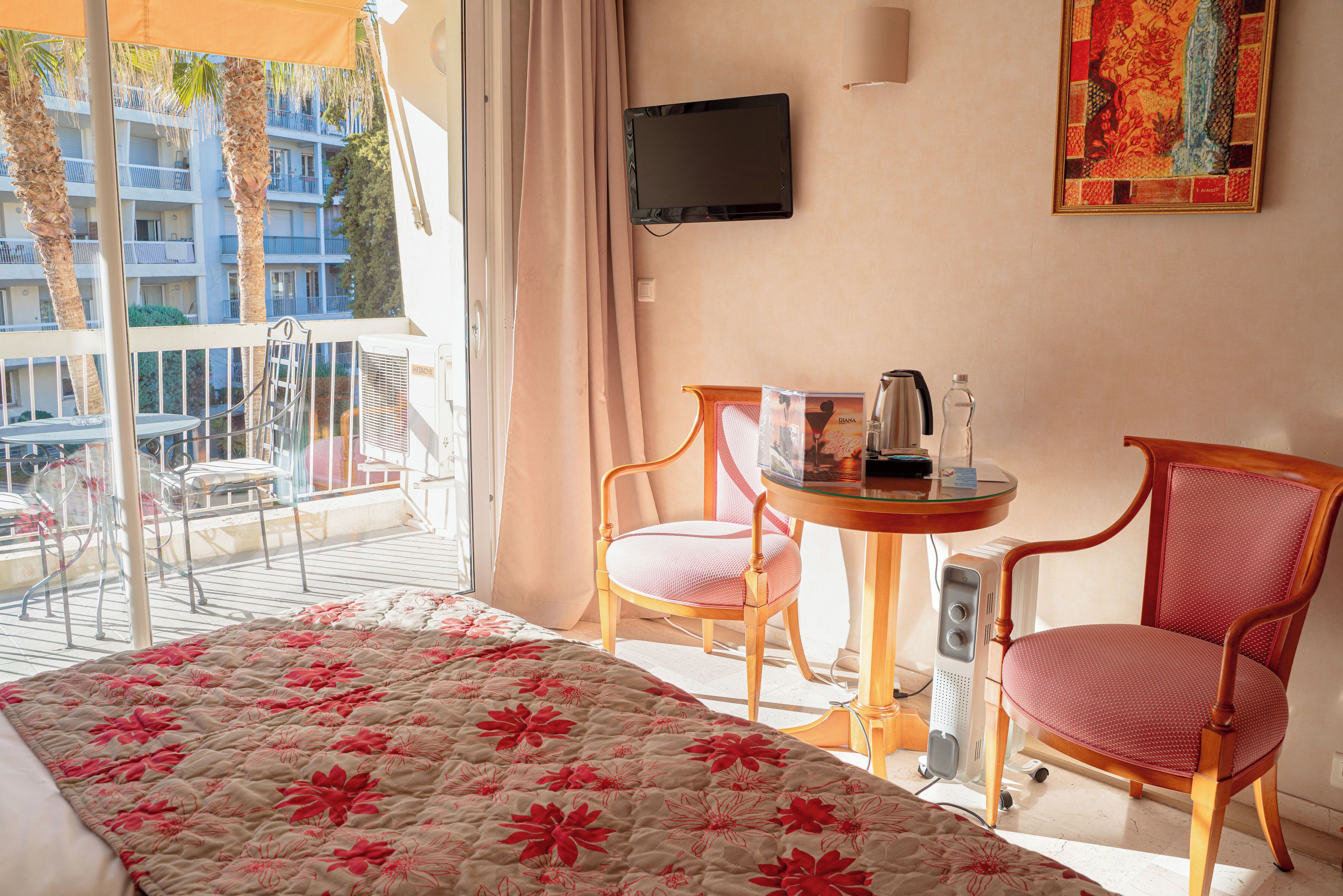 A hotel room with a balcony, a bed with a floral bedspread, two chairs, a small table with a coffee pot, and a television on the wall.