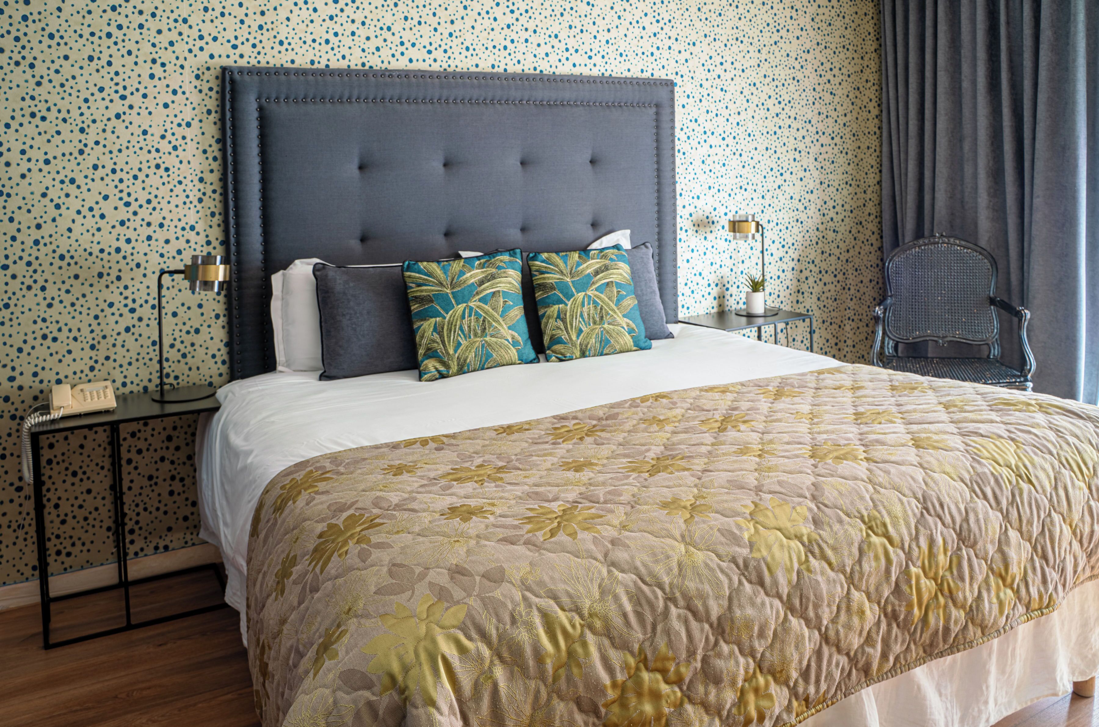 Cama bem arrumada com cabeceira estofada, duas almofadas decorativas, mesa de cabeceira com abajur e uma cadeira.