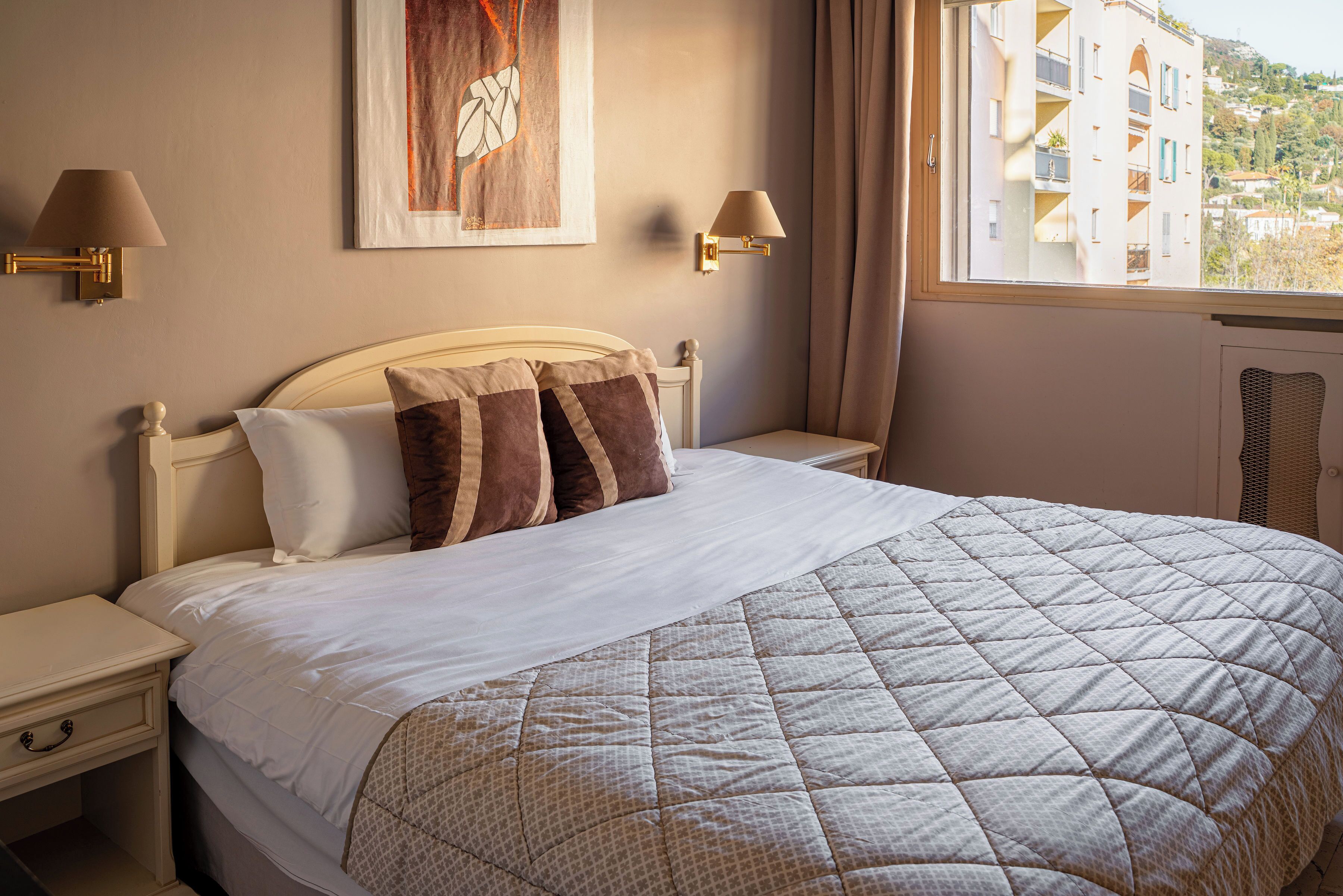 A neatly made bed with a quilted bedspread, two bedside lamps, a framed artwork above the bed, and a window with curtains.