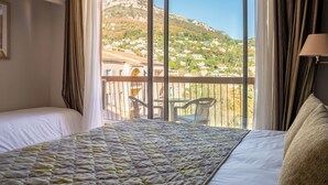 A hotel room with a large bed, a balcony with a view of a hillside town, and a table with chairs outside. - Hotel Diana (Vence)