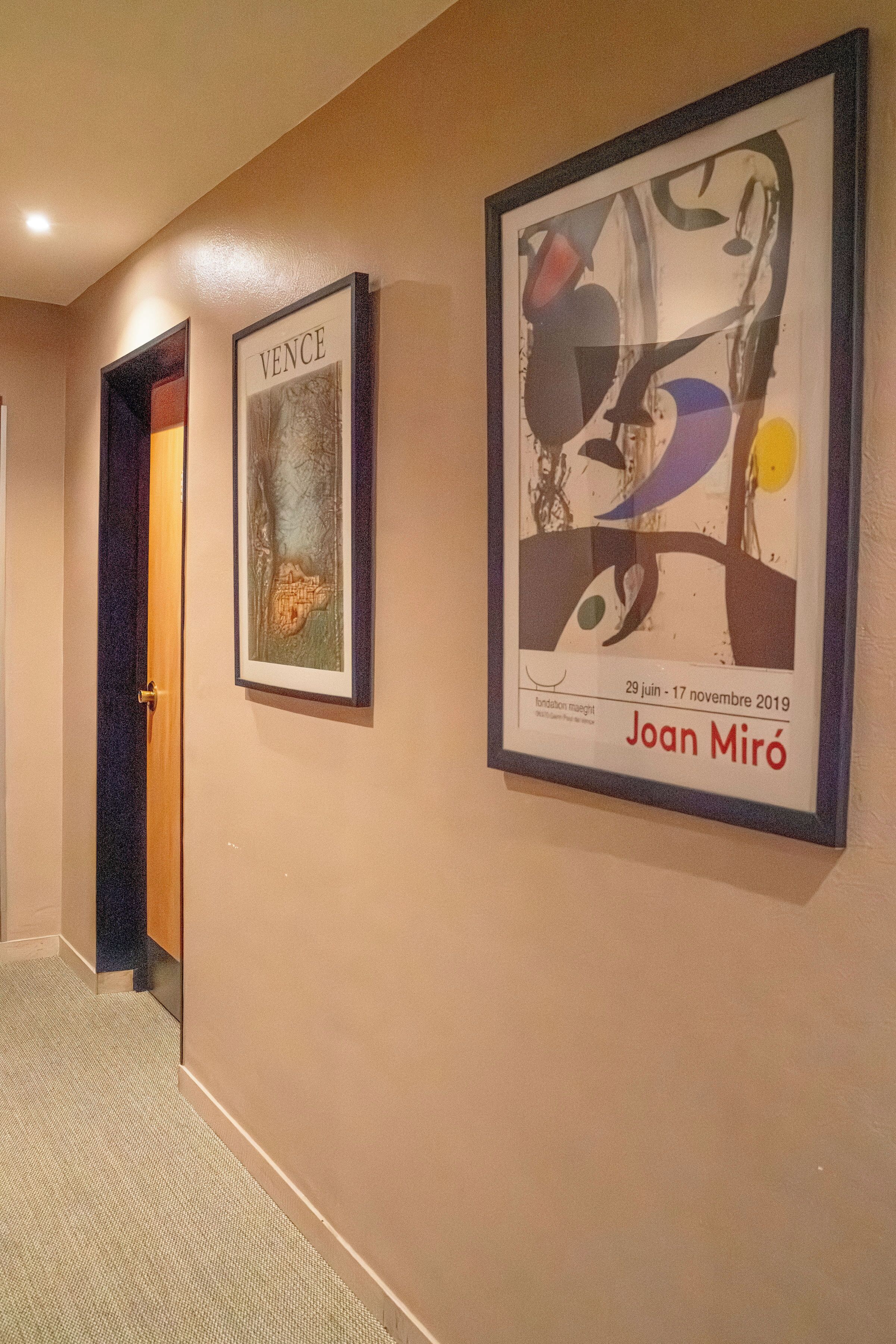 A hallway with two framed artworks on the wall, a door to a room, and a carpeted floor.