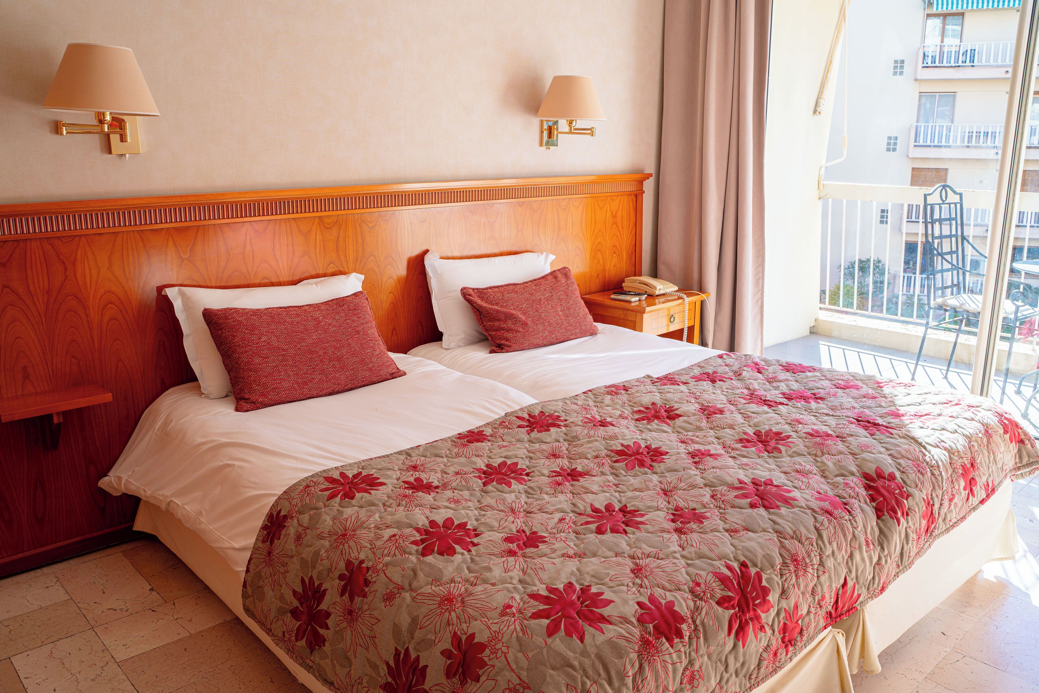 Habitación de hotel con cabecera de madera, dos camas con ropa de cama roja y blanca, colcha de estampado floral, dos lámparas de noche, una mesita de noche con teléfono y vistas a un balcón con barandilla.