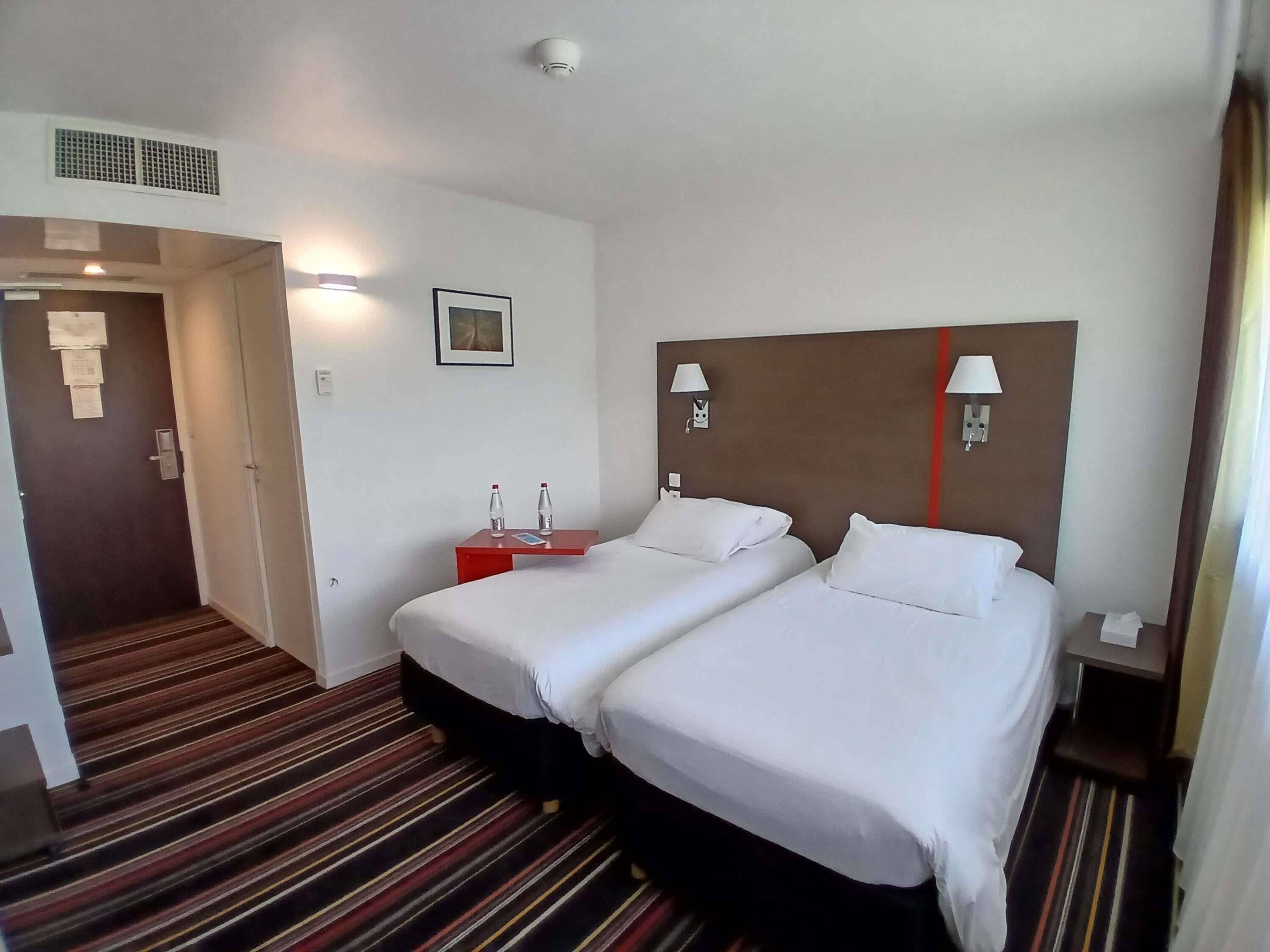 A hotel room with two beds, a red nightstand, a wall-mounted lamp, and a door leading to another room.