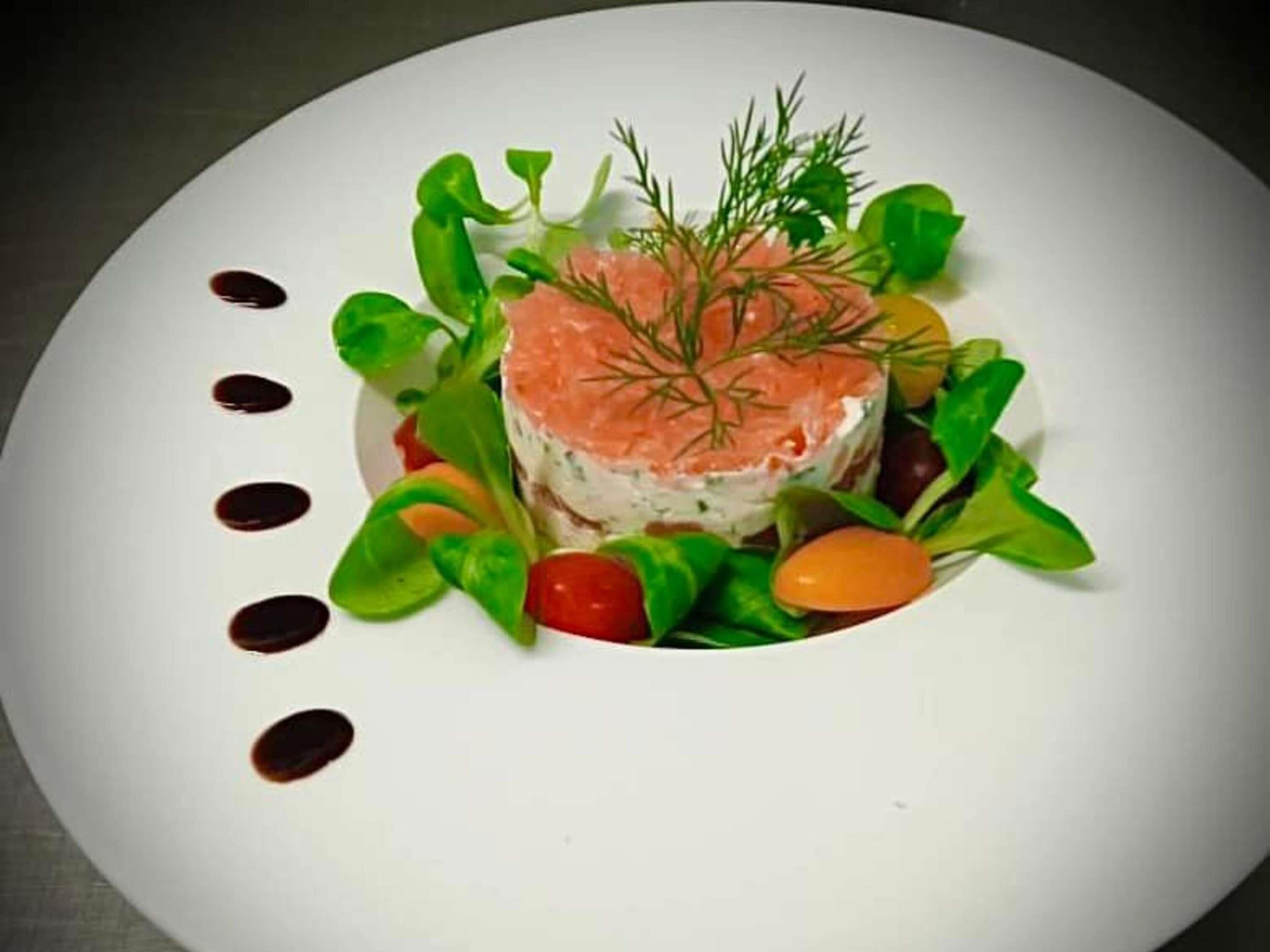 Une assiette blanche sur laquelle est disposée une tranche de saumon au centre, entourée de légumes verts et de tomates cerises, ainsi que cinq petites gouttes de sauce foncée sur le bord de l’assiette.