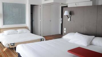 Habitación de hotel con dos camas, una mesita de noche roja, una ventana con persianas, una puerta y una lámpara fijada a la pared.