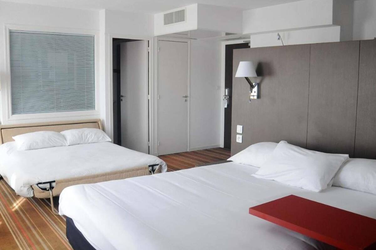 A hotel room with two beds, a red side table, a window with blinds, a wall-mounted lamp, and a door leading to another room.
