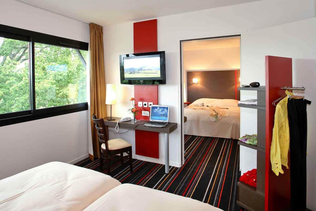 A hotel room with a bed, a desk with a computer, a TV, and a view of trees through the window.