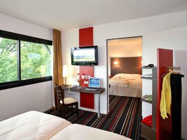 Habitación de hotel con cama, escritorio con computadora, televisor y vista a árboles por la ventana.