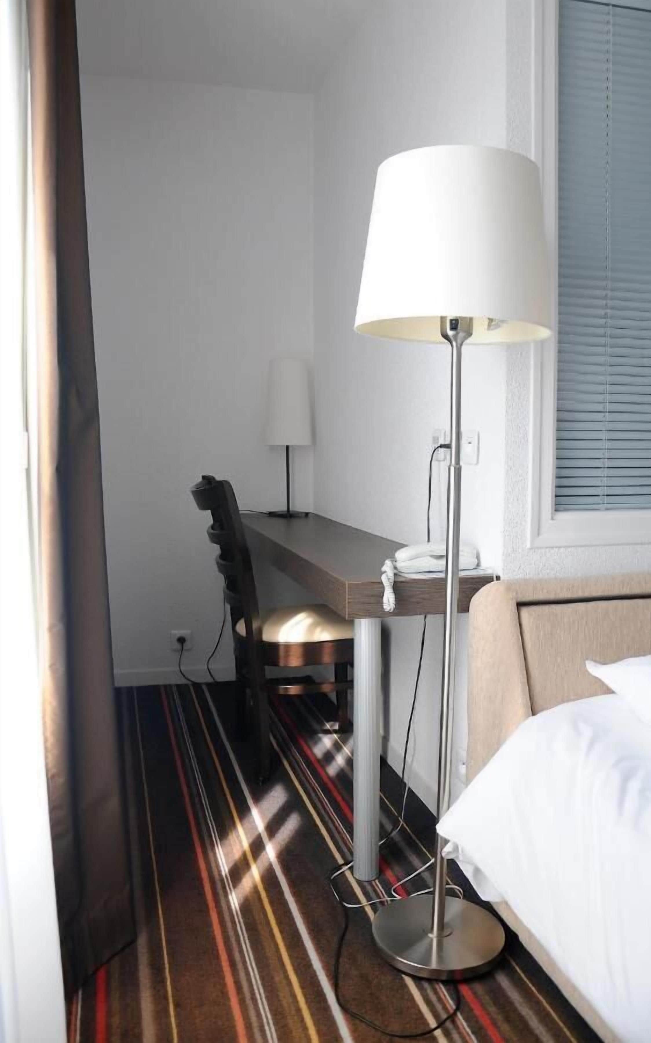Une chambre d’hôtel avec un lit, un bureau, une chaise, une lampe et un tapis à rayures.
