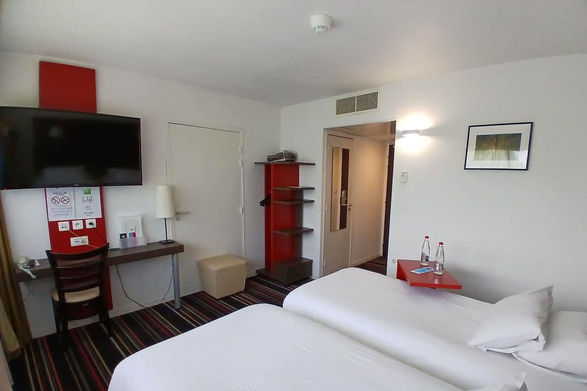 A hotel room with a bed, a desk with a chair, a television, a red cabinet, a small stool, and a framed picture on the wall.