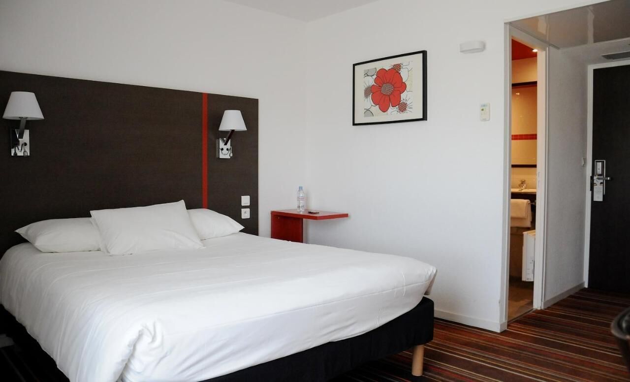 Habitación de hotel con una cama grande, dos lámparas de noche, una mesita con una botella de agua y una puerta que da a un baño.