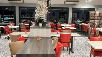 Een restaurant met een lange centrale tafel, rode en beige stoelen en een decoratieve witte bloemstelling.