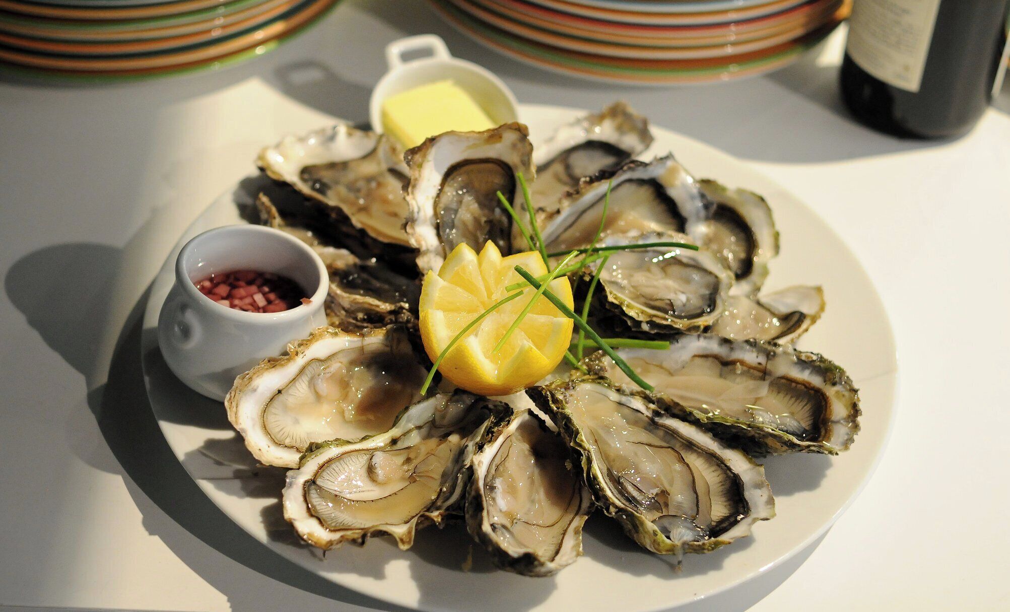 Un plato de ostras con gajos de limón y cebollino, acompañado de un recipiente pequeño con salsa roja y un trozo de mantequilla.