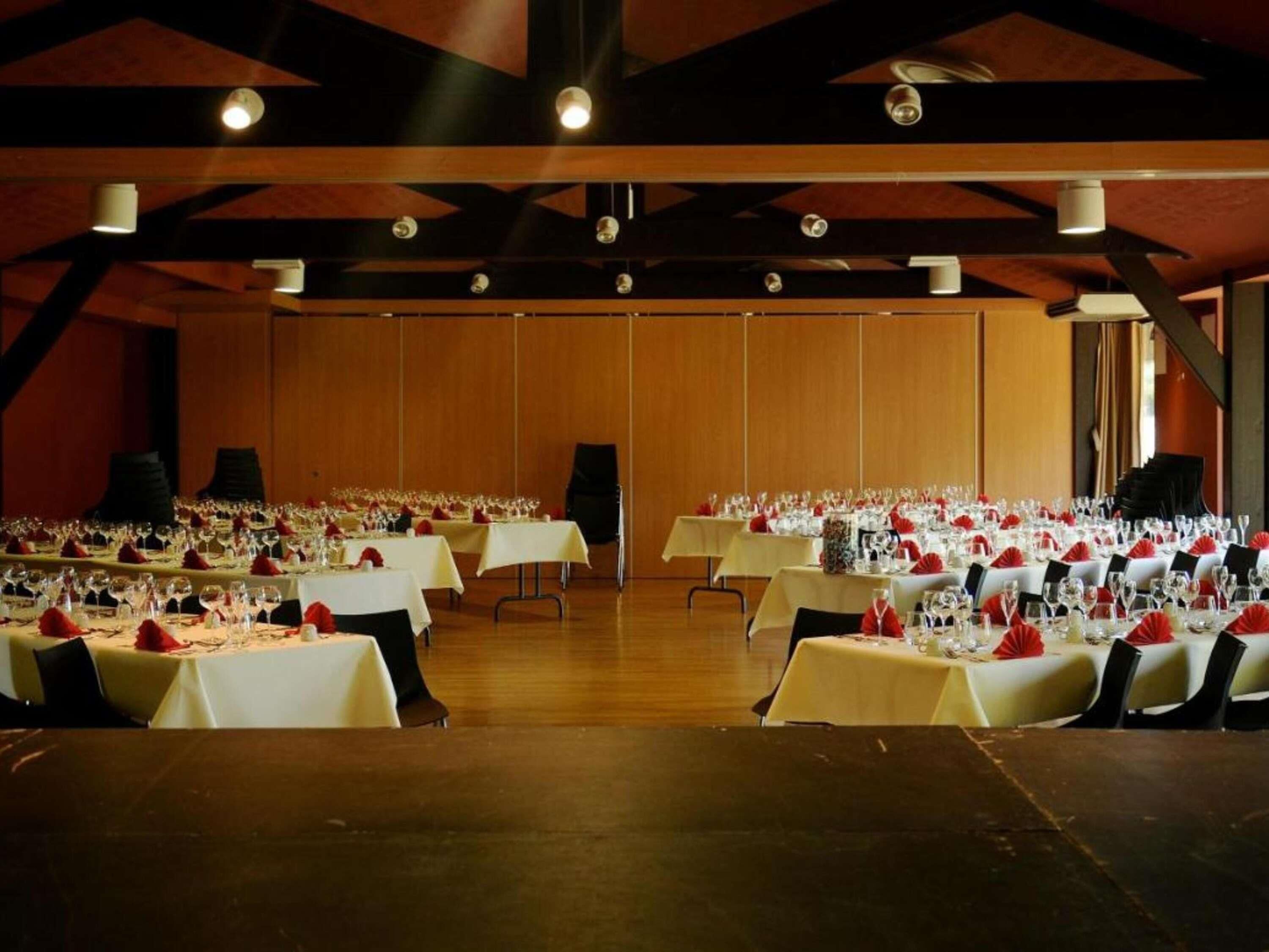 A banquet hall with multiple tables set for a formal event, featuring a high ceiling with exposed beams and a wooden floor.