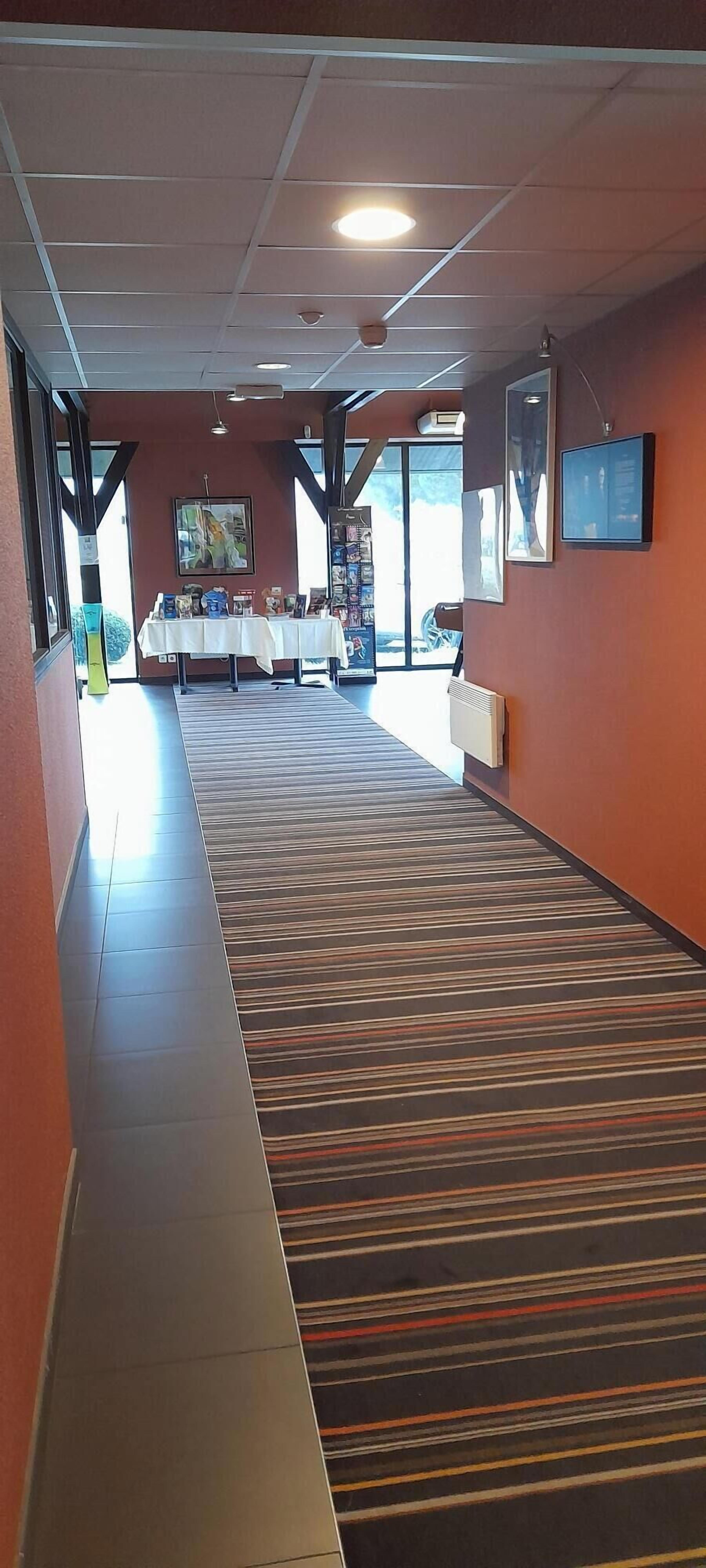 Un pasillo de hotel con alfombra a rayas, un mostrador de recepción, cuadros enmarcados en la pared y una mesa con folletos.