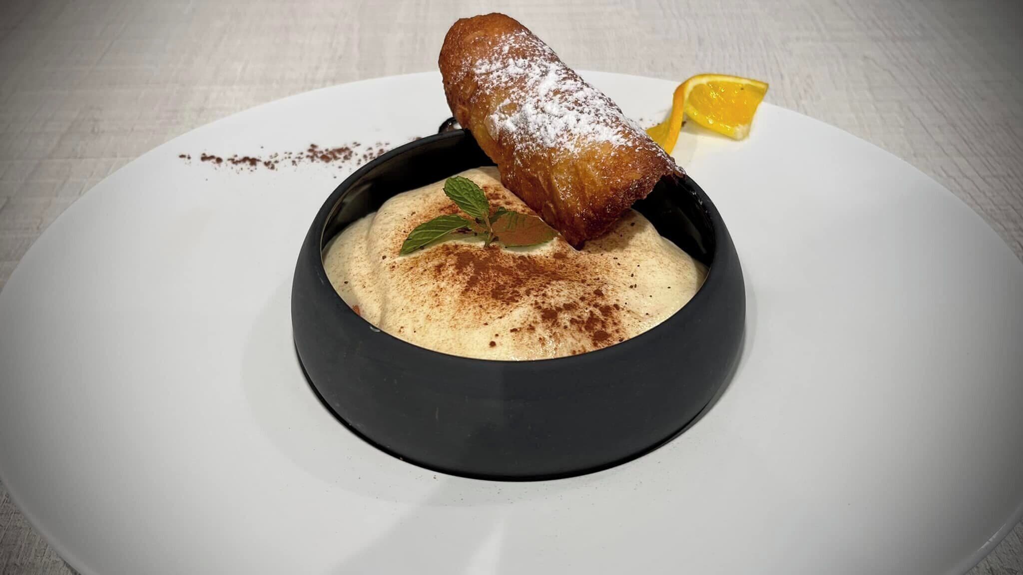 A dessert in a black bowl on a white plate, garnished with a powdered topping and a lemon slice.