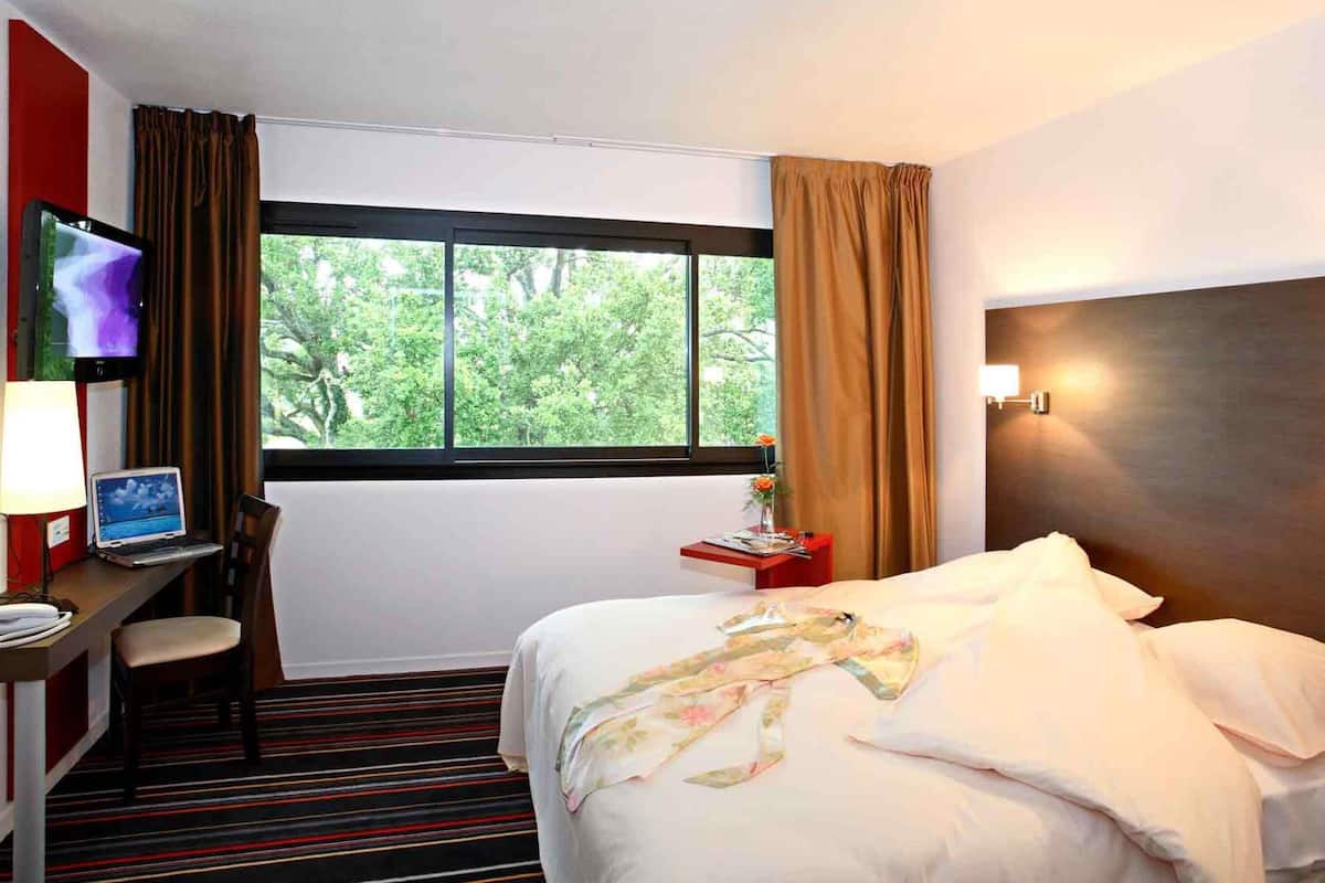 A hotel room with a bed, a desk with a laptop, a TV, and a window with curtains.