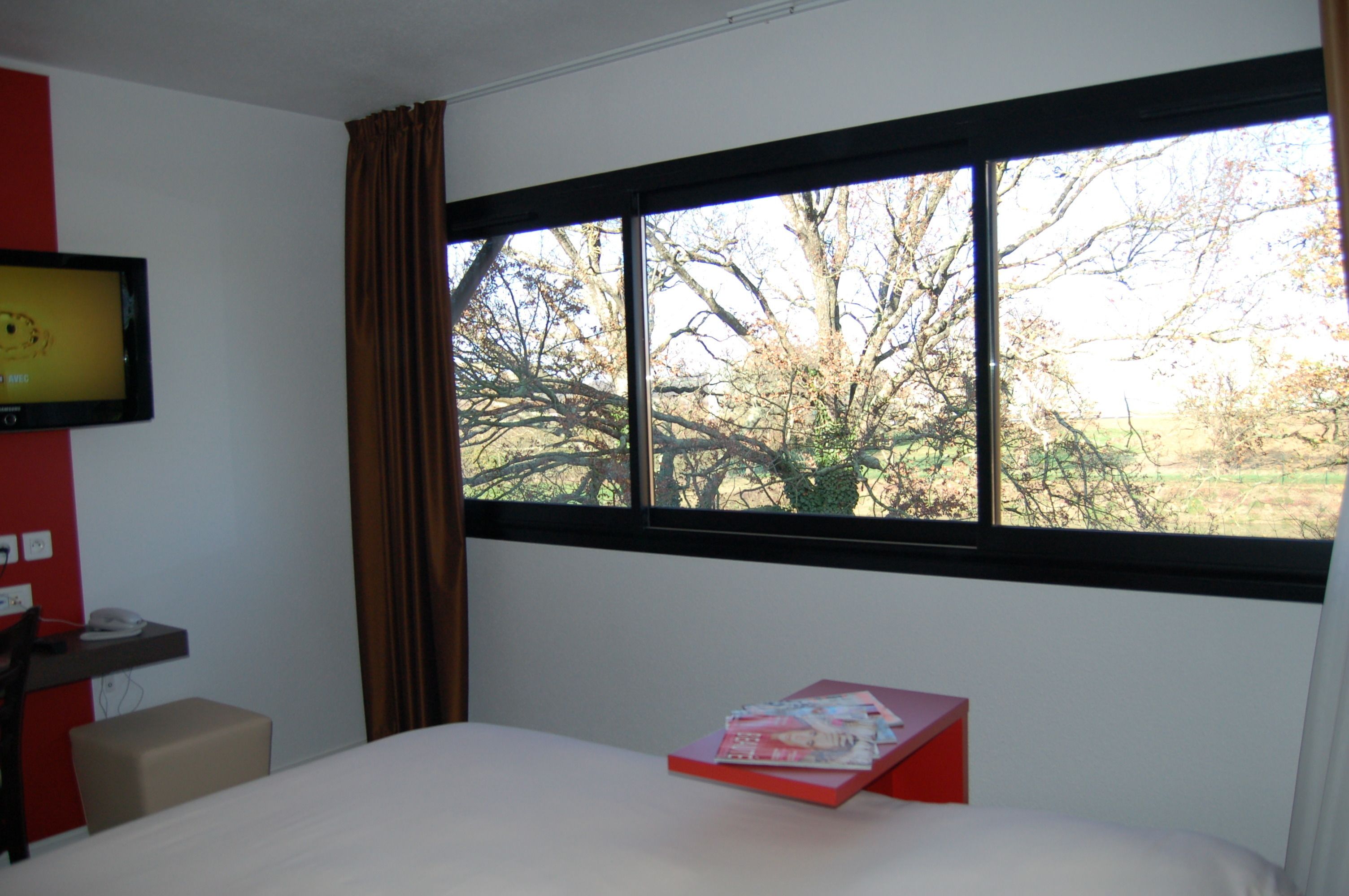 A hotel room with a large window, a bed, a desk with a chair, and a television on the wall.