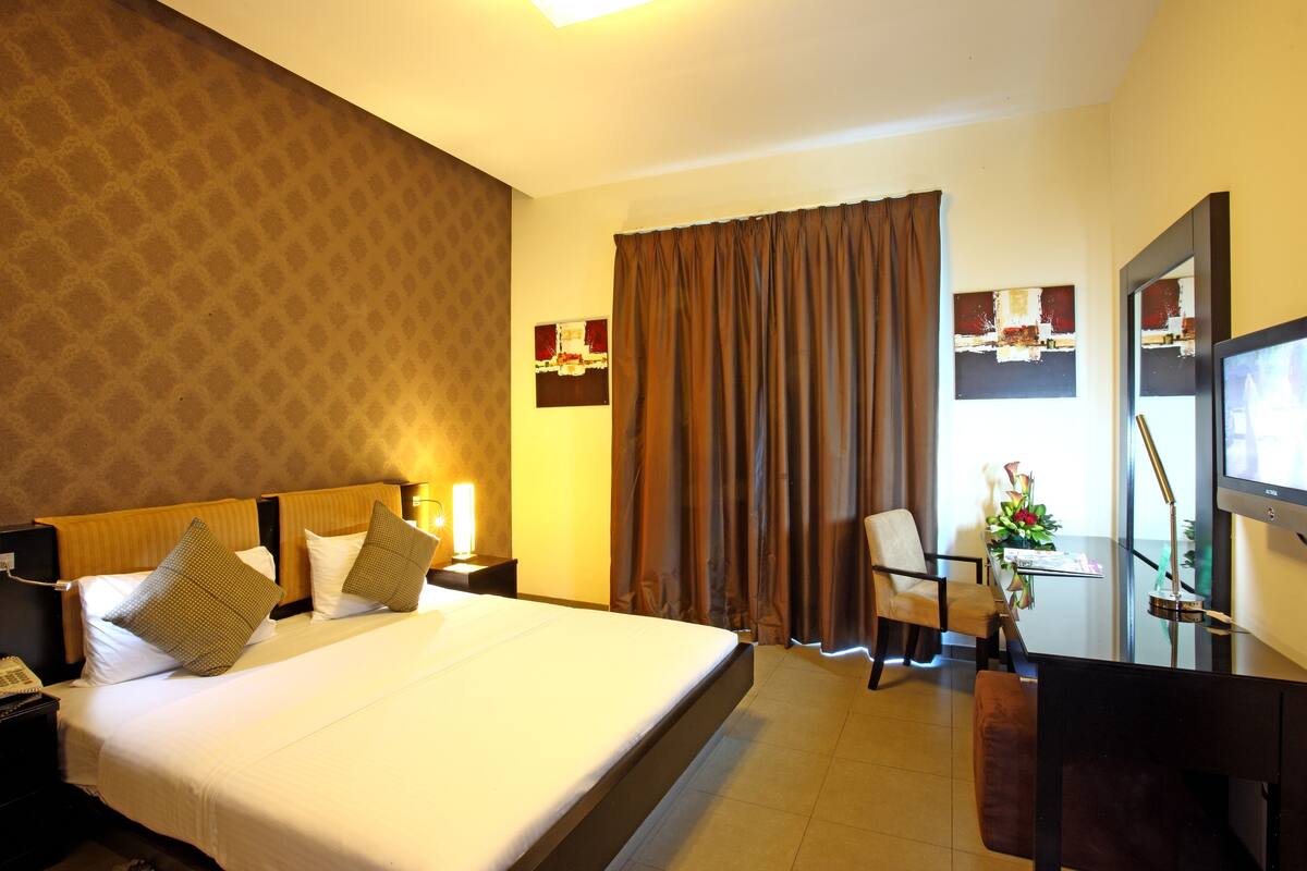 Une chambre d’hôtel avec un grand lit, un bureau avec une chaise, une télévision et une fenêtre avec des rideaux.