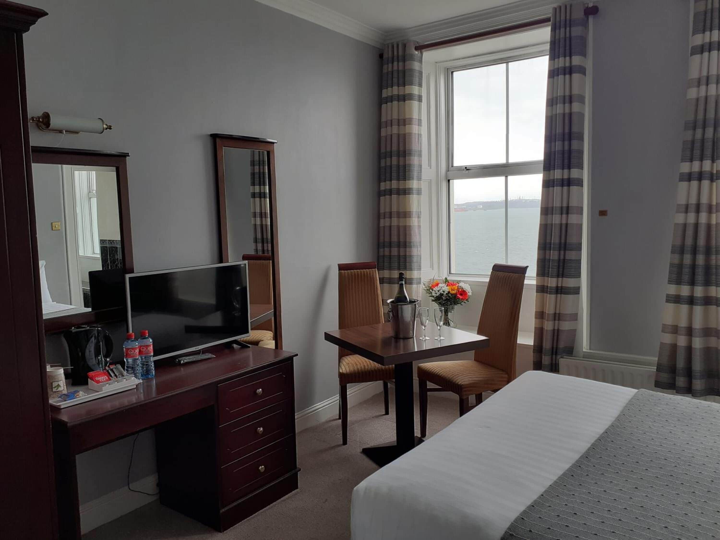 A hotel room with a bed, a desk with a TV, a mirror, a chair, and a window with curtains.