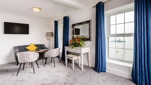 A hotel room with a grey sofa, two chairs, a white desk, a mirror, and a large window with blue curtains. - Commodore Hotel (Cobh)