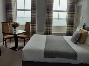 A hotel room with a bed, a dining table with chairs, and a view of the sea through large windows.