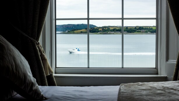 A bedroom with a large window overlooking a body of water and distant hills. - Commodore Hotel (Cobh)