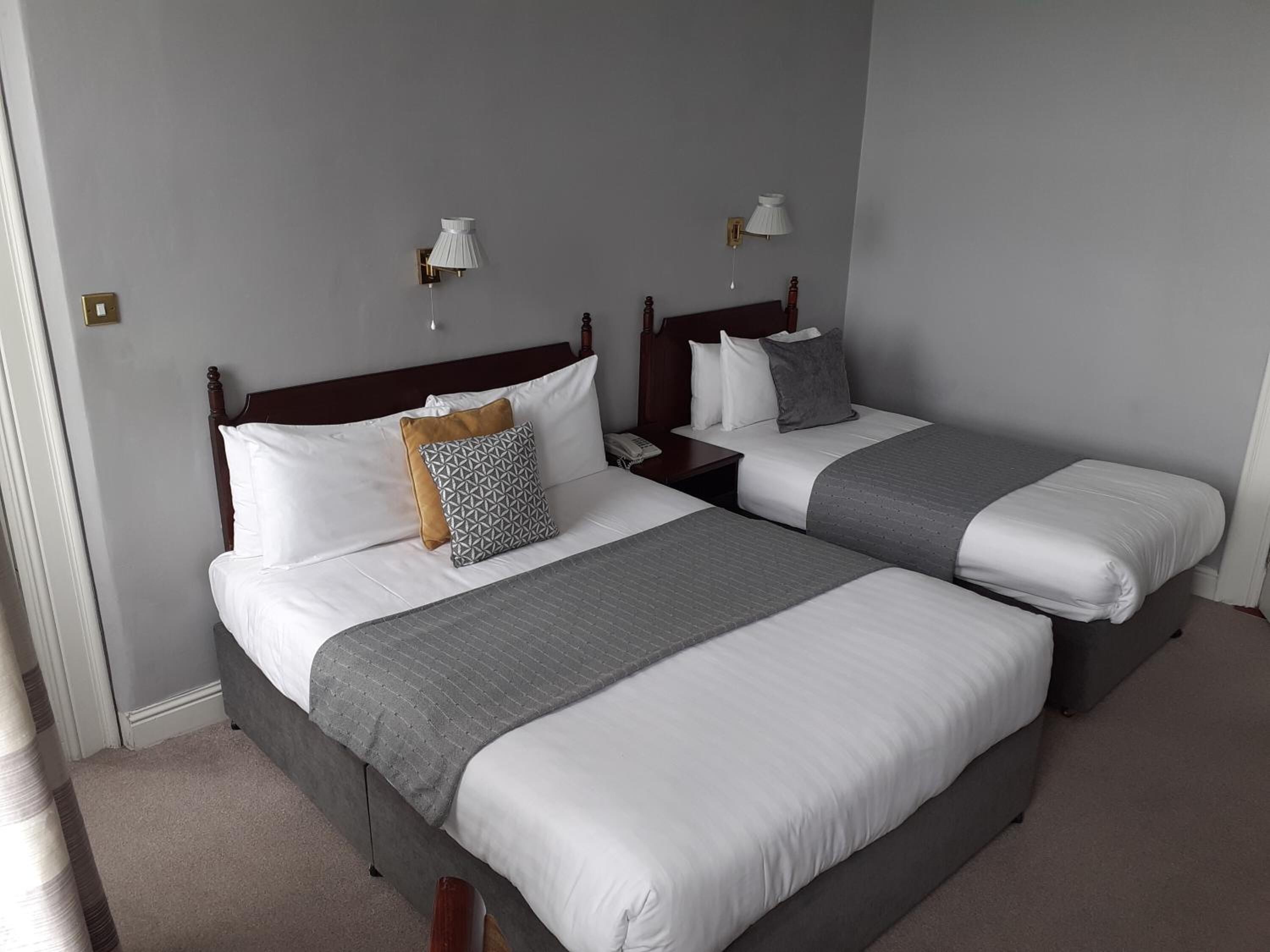 A hotel room with two beds, each with a wooden headboard, white bedding, and a grey blanket. There are bedside tables with lamps and a telephone on one of the beds.