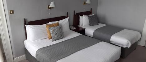 A hotel room with two beds, each with a wooden headboard, white bedding, and a grey blanket. There are bedside tables with lamps and a telephone on one of the beds.