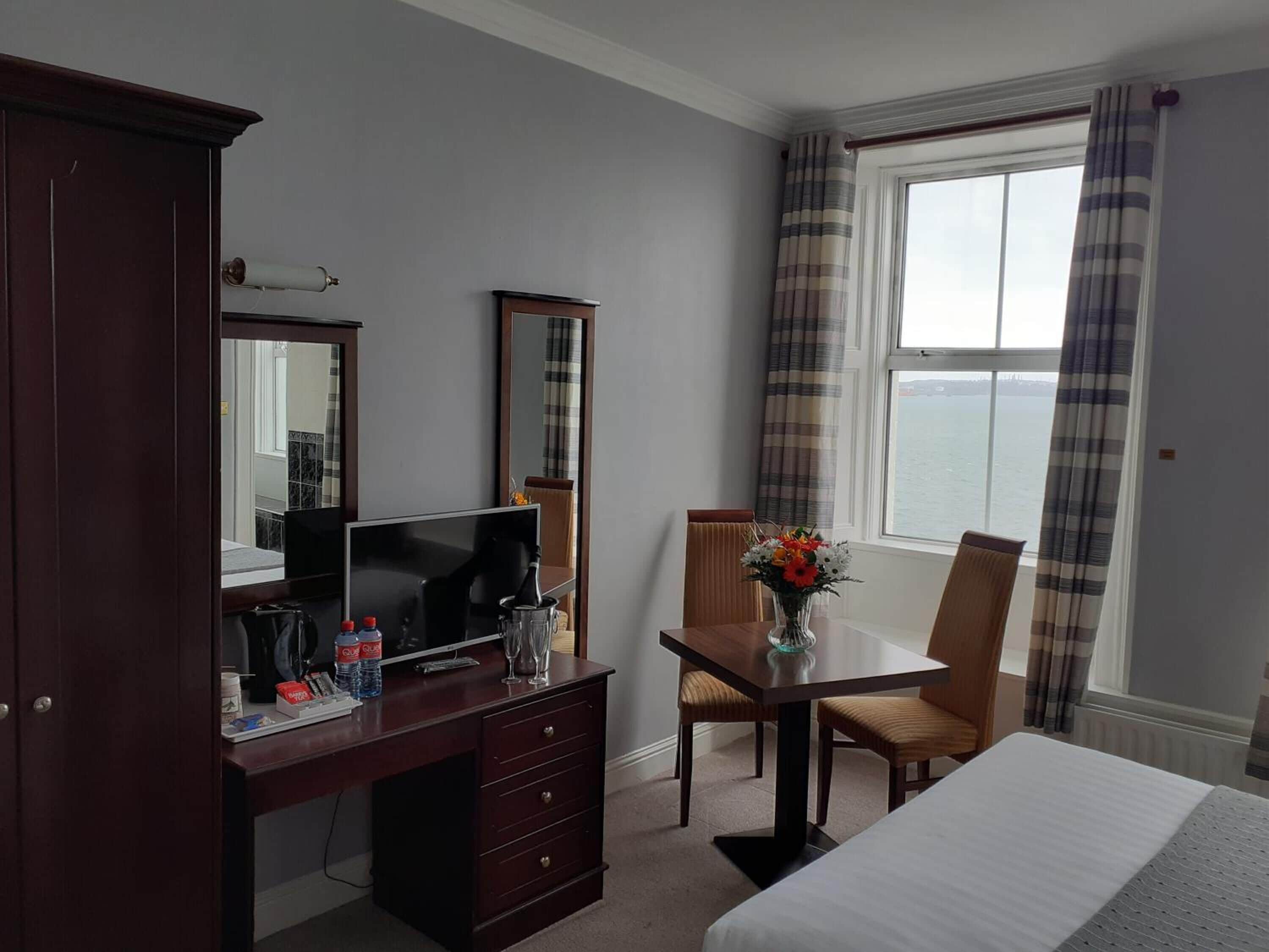 A hotel room with a bed, a desk with a TV, a mirror, a chair, a vase with flowers, and a window with curtains.