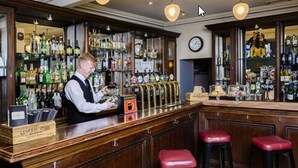 A bar with a bartender, a beer tap, and a shelf with various bottles. - Commodore Hotel (Cobh)