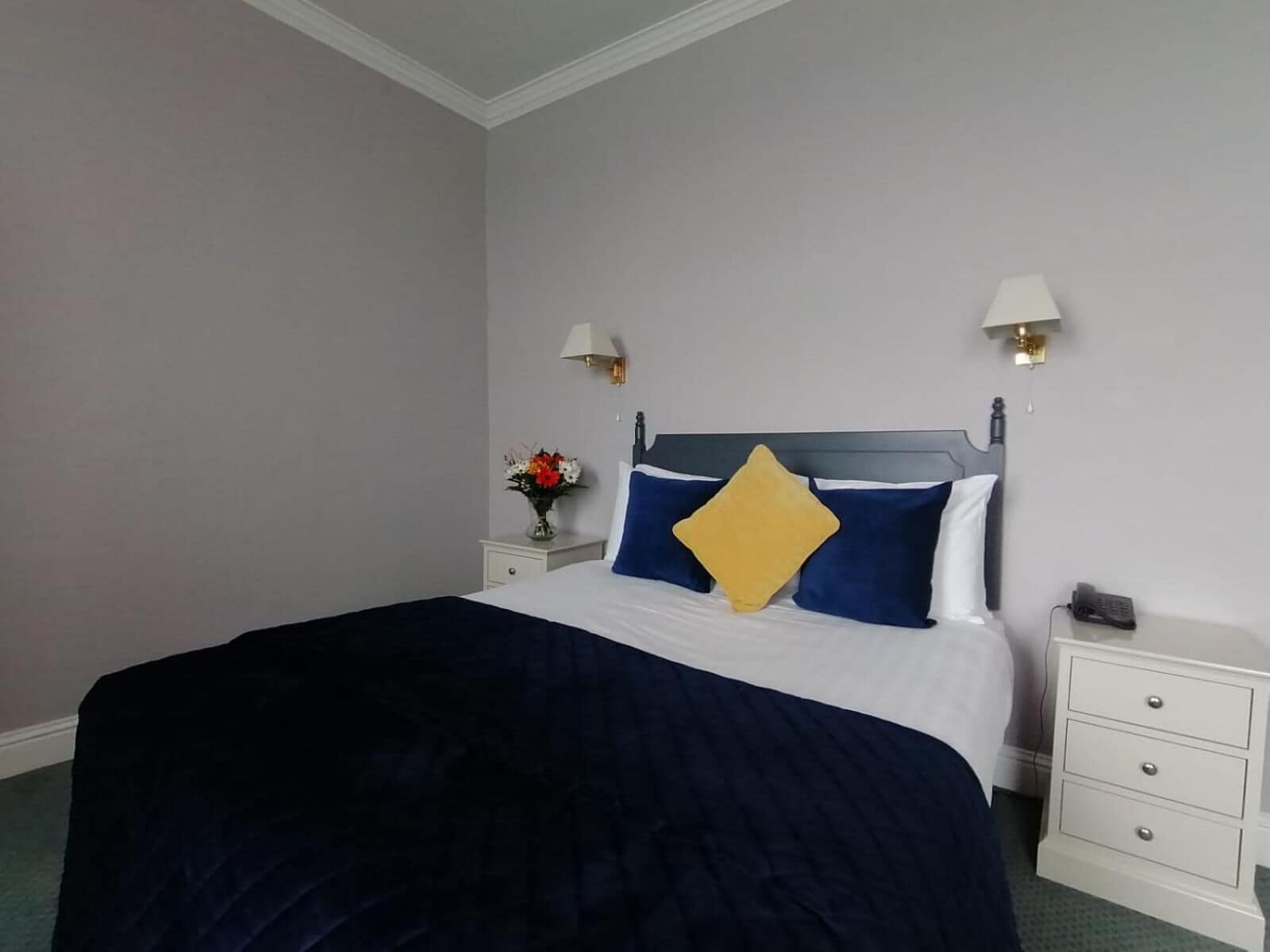 A neatly made bed with a dark blue comforter and a single yellow pillow. There are two bedside tables with lamps and a vase of flowers.