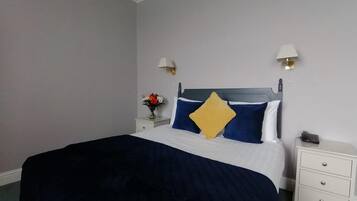 A neatly made bed with a dark blue comforter and a single yellow pillow. There are two bedside tables with lamps and a vase of flowers.