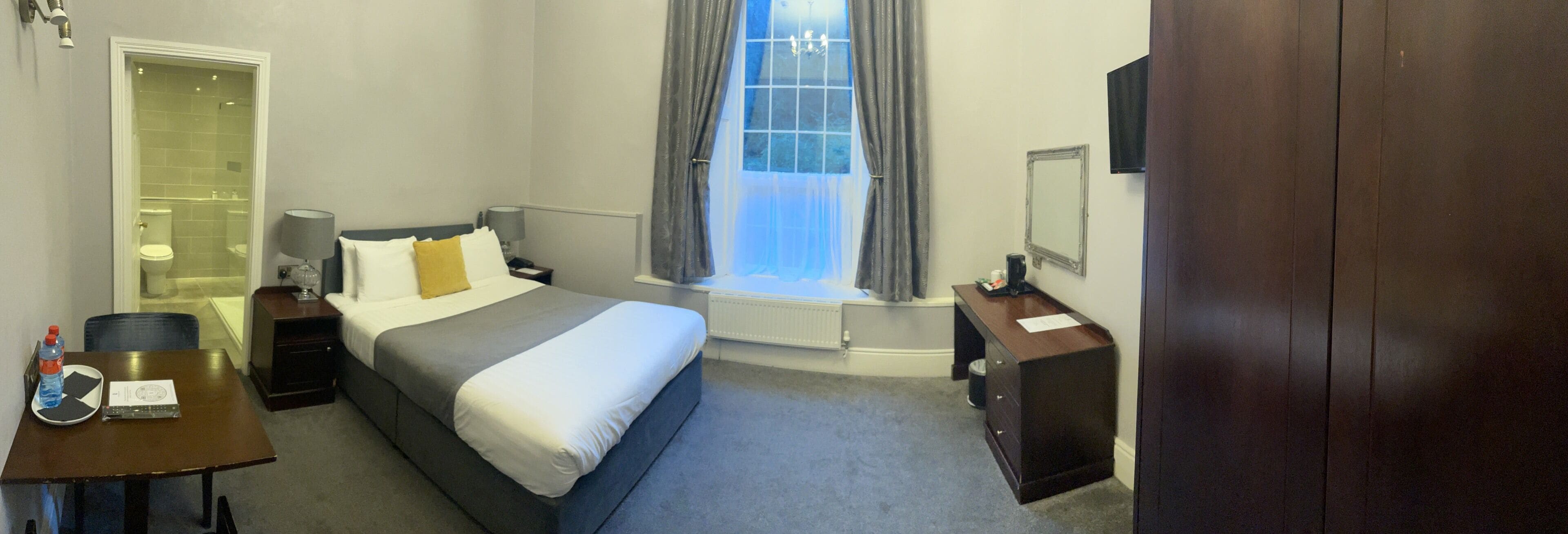 A hotel room with a bed, bedside tables, a desk, a chair, a window with curtains, a radiator, and a TV.